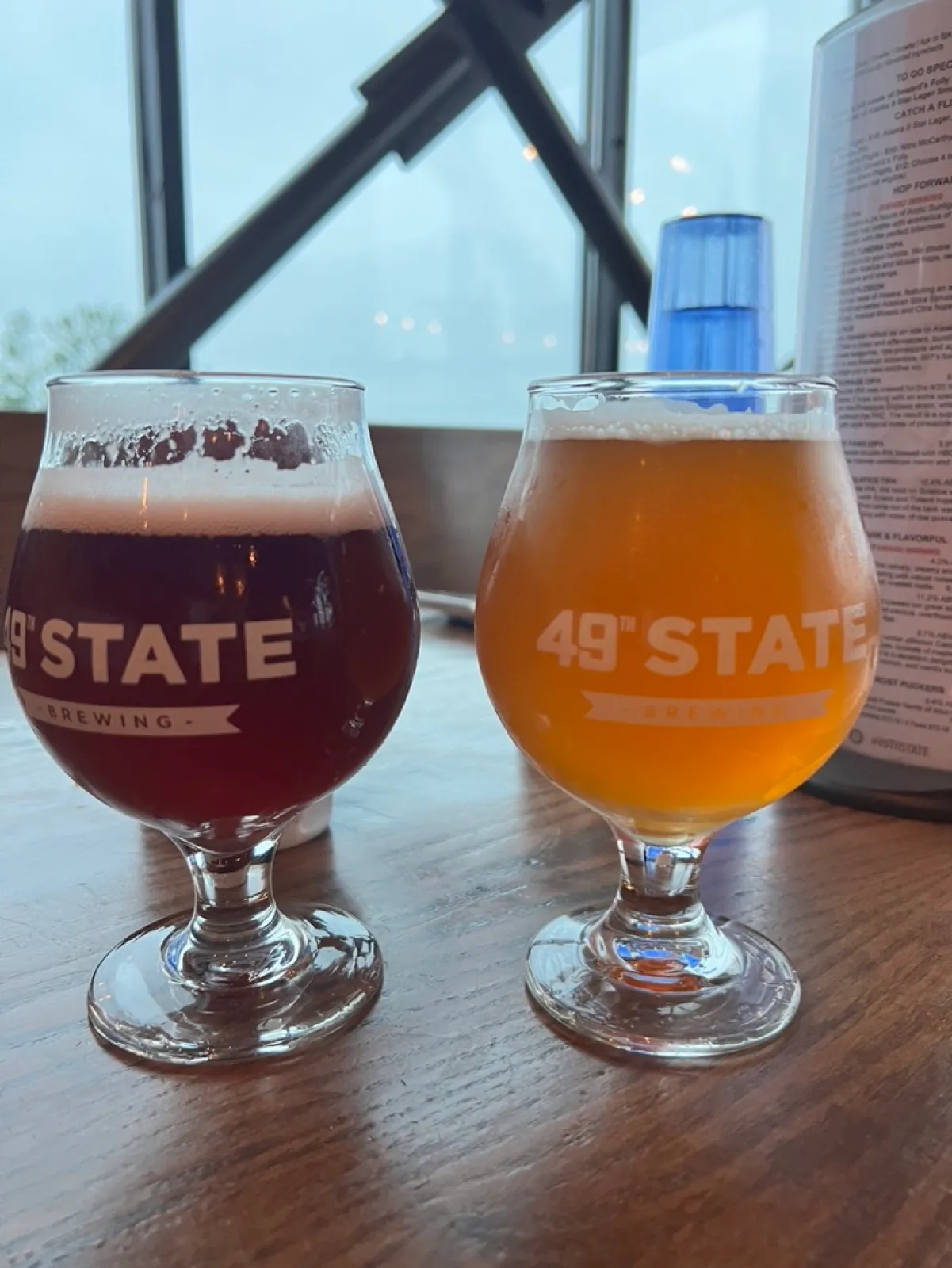 Two snifter glasses on a wooden table, both labeled '49th State Brewing' — one amber, one hazy gold — with a can and menu in soft  focus behind.