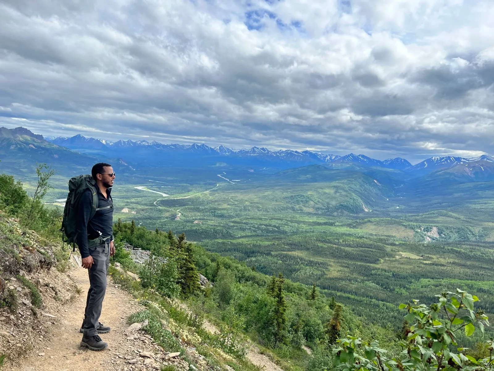 Robel on a thin ridge trail with a backpack, looking out over a wide green river valley and snow-streaked mountains under a cloudy  sky.