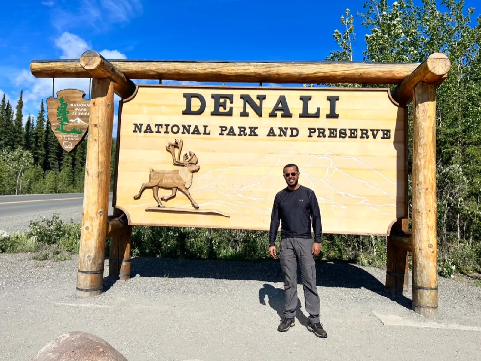 Robel standing in front of the wooden Denali National Park and Preserve entrance sign with a carved caribou on a clear-sky day.