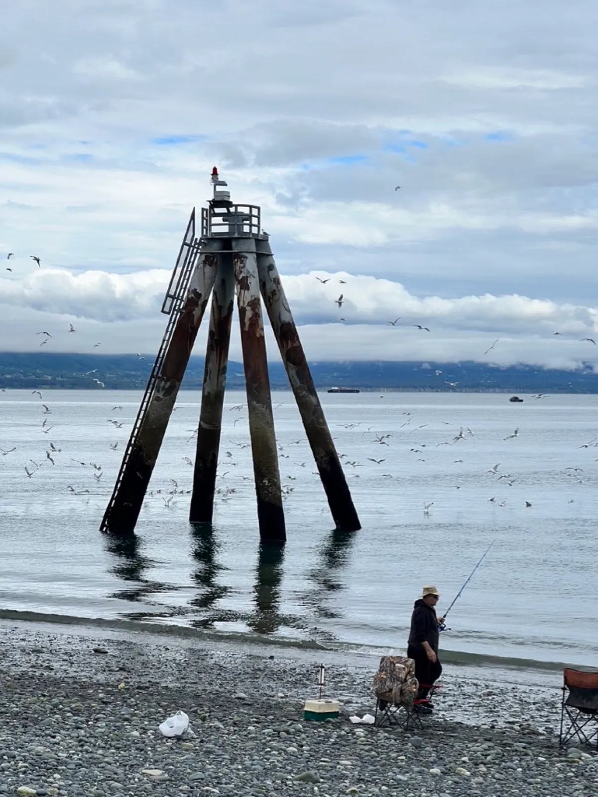 A weathered four-legged fishing tower in shallow water with seagulls circling and an angler casting from the rocky beach.
