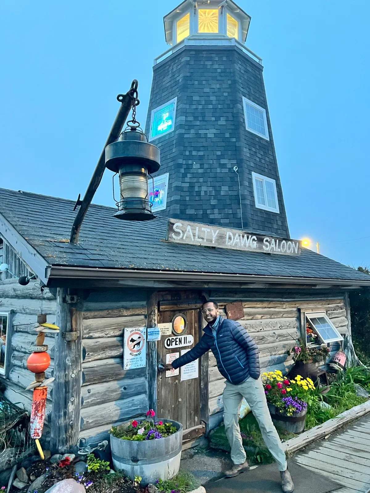 The Salty Dawg Saloon — a log cabin with a tall slate-shingled lighthouse tower — at dusk, with flowers along the entrance.