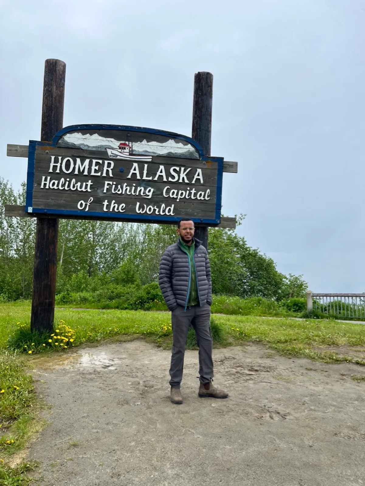 Robel standing in front of the Homer, Alaska 'Halibut Fishing Capital of the World' sign on a cloudy day.