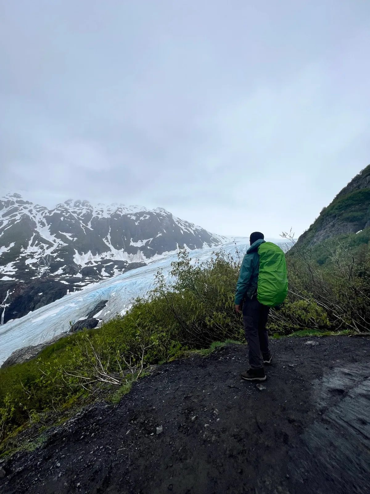 Robel in a green pack-cover standing on a dark dirt trail, looking out at the blue tongue of Exit Glacier flowing down a  mountainside.