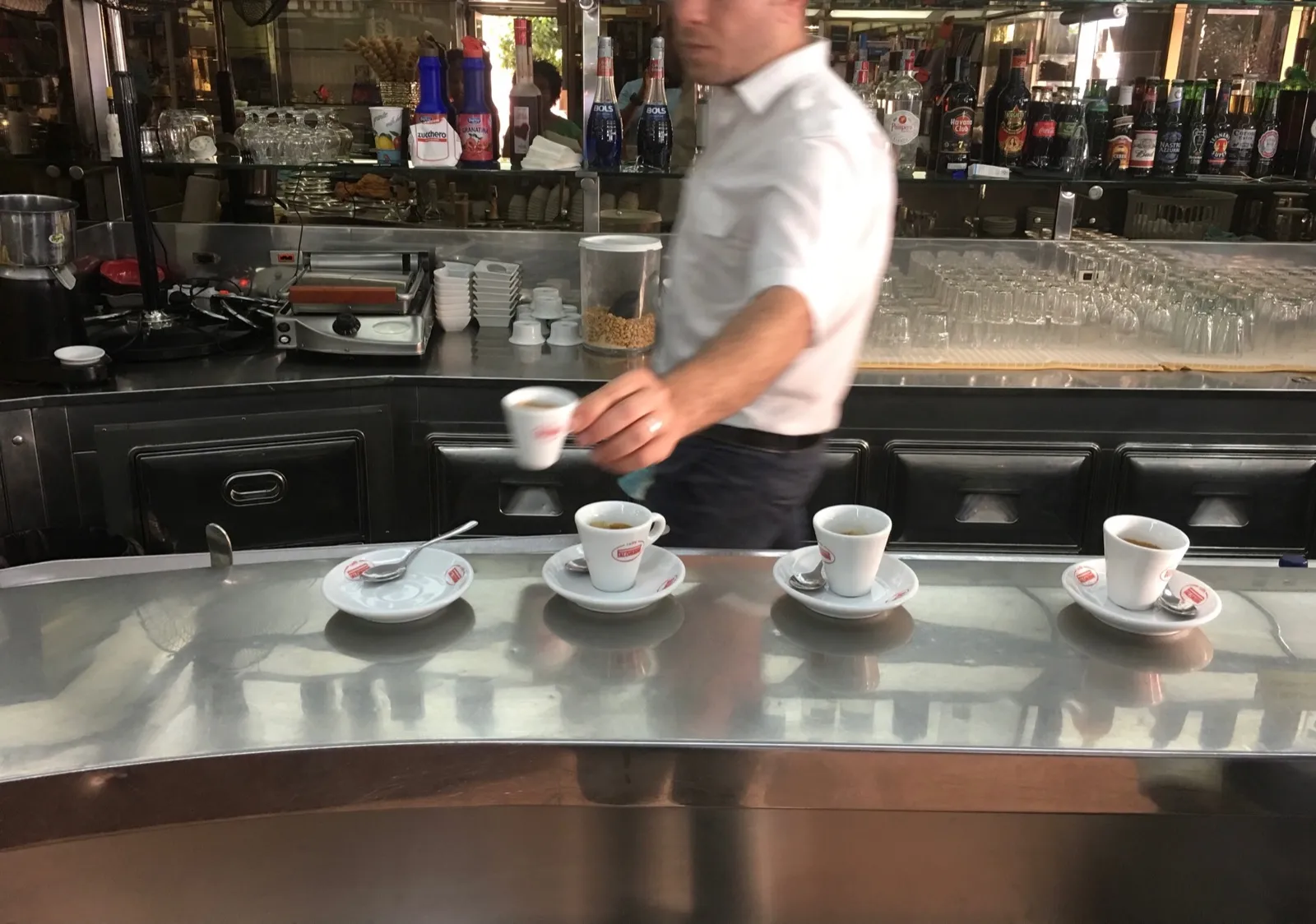 Four small espresso cups on saucers lined up along a polished stainless-steel bar, with a barista in a white shirt placing the  fourth, surrounded by bottles and glassware.