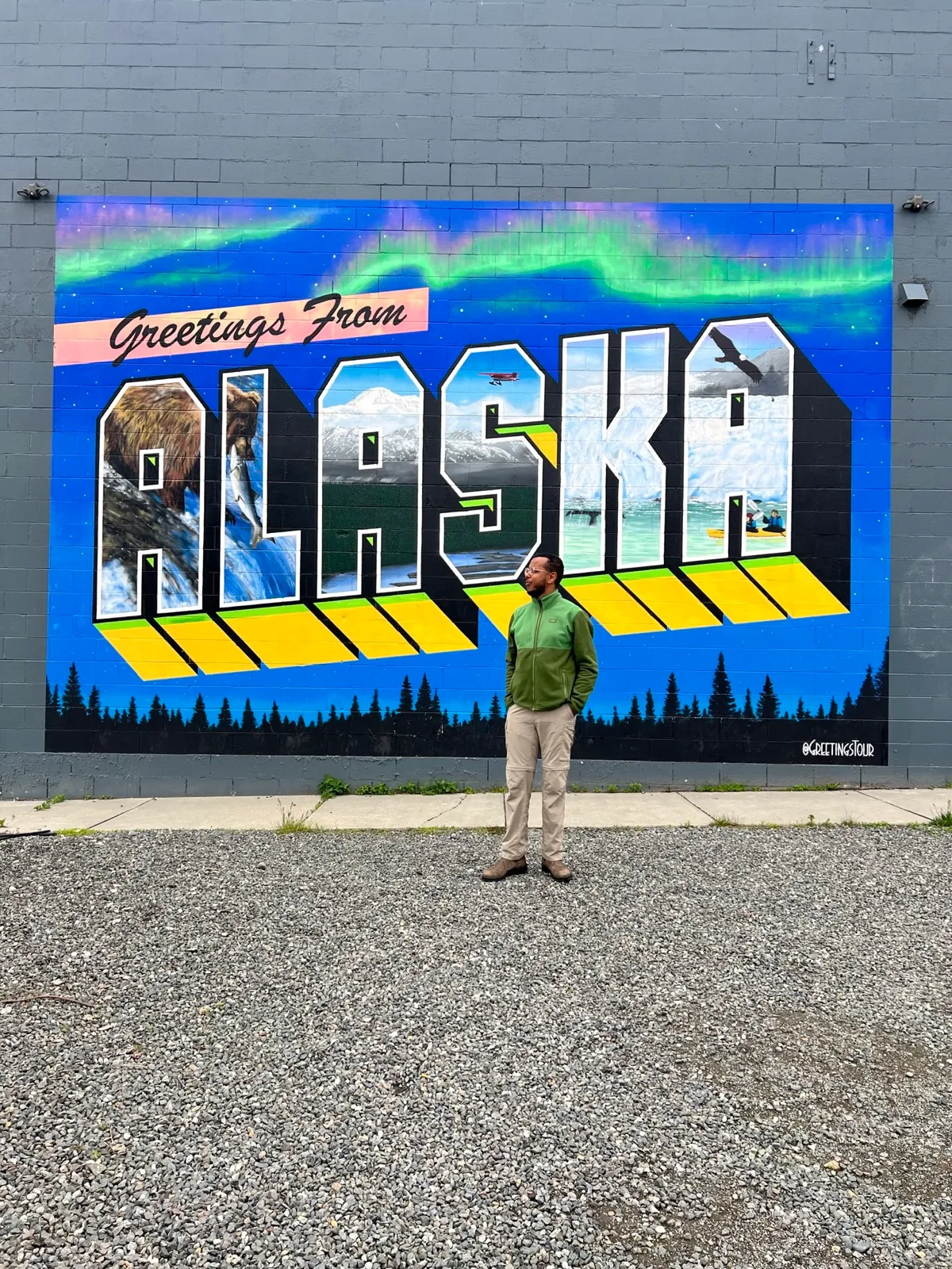 Robel standing in front of a large 'Greetings From Alaska' postcard-style mural with a grizzly bear, glacier, and the northern  lights.