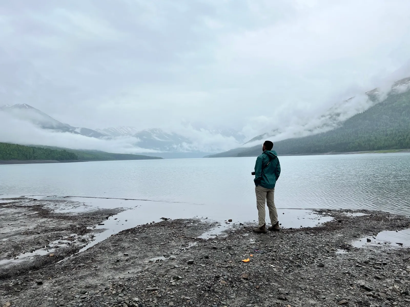 Robel in a green rain shell standing on the pebbled shore of Eklutna Lake, with low clouds wrapped around the surrounding ridges.