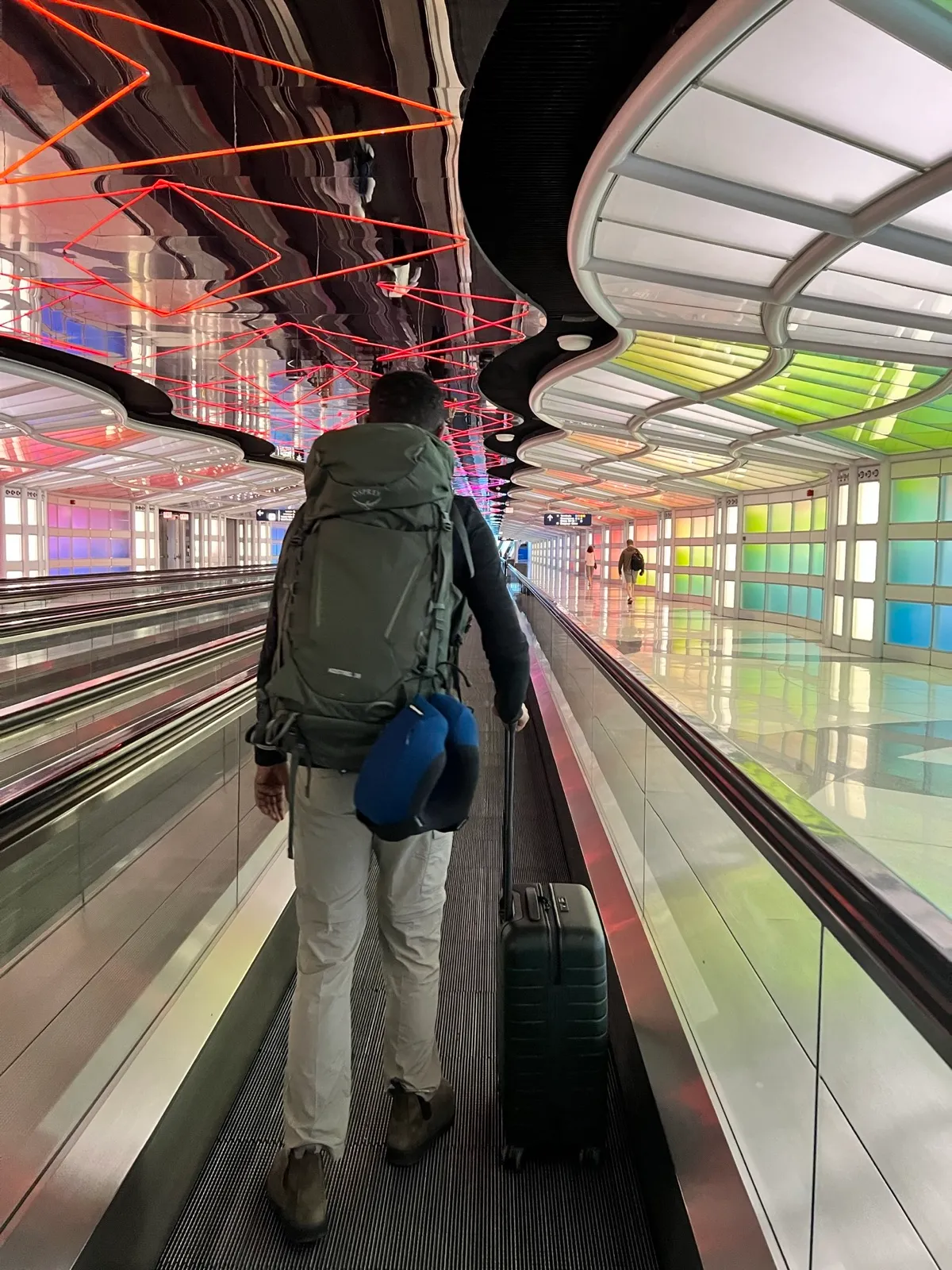 Robel walking a moving walkway in Chicago O'Hare's underground tunnel with a luggage trolley, the ceiling lit by changing neon  colors.