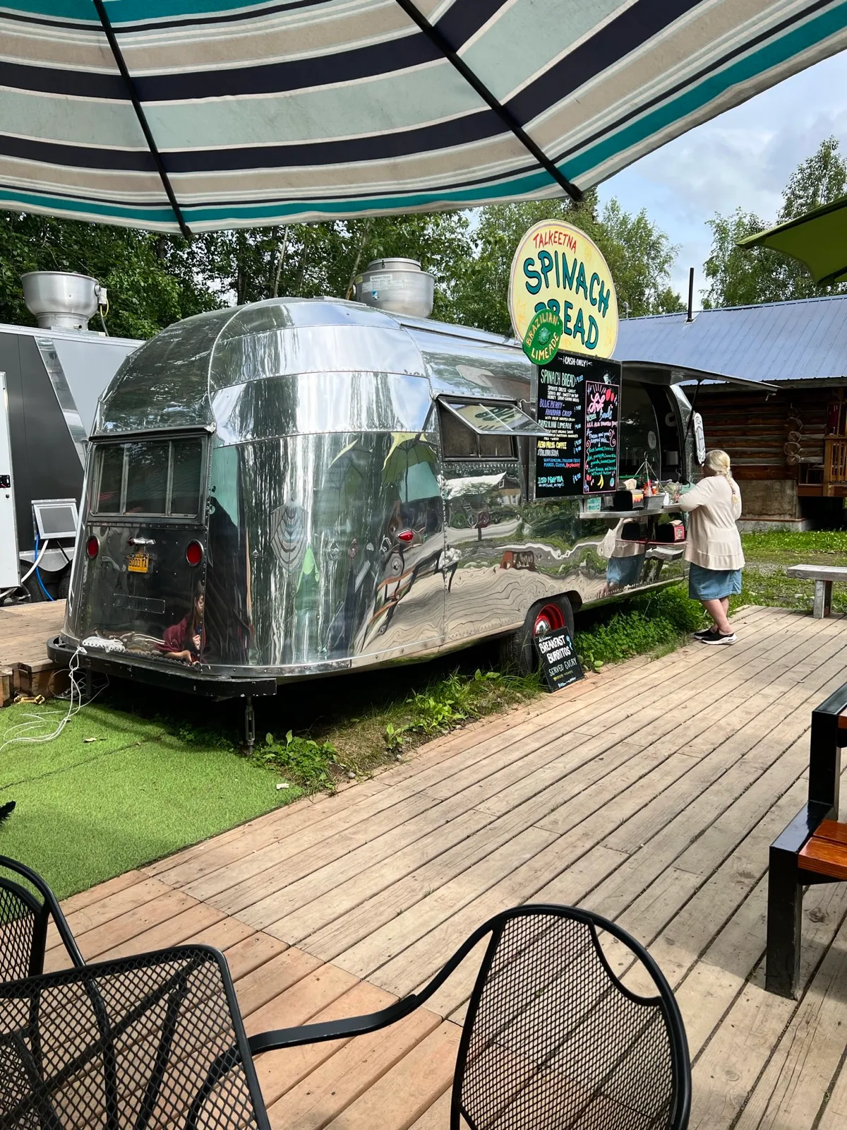 A polished silver Airstream food trailer in Talkeetna selling Brazilian limeade and spinach bread, under a striped umbrella.