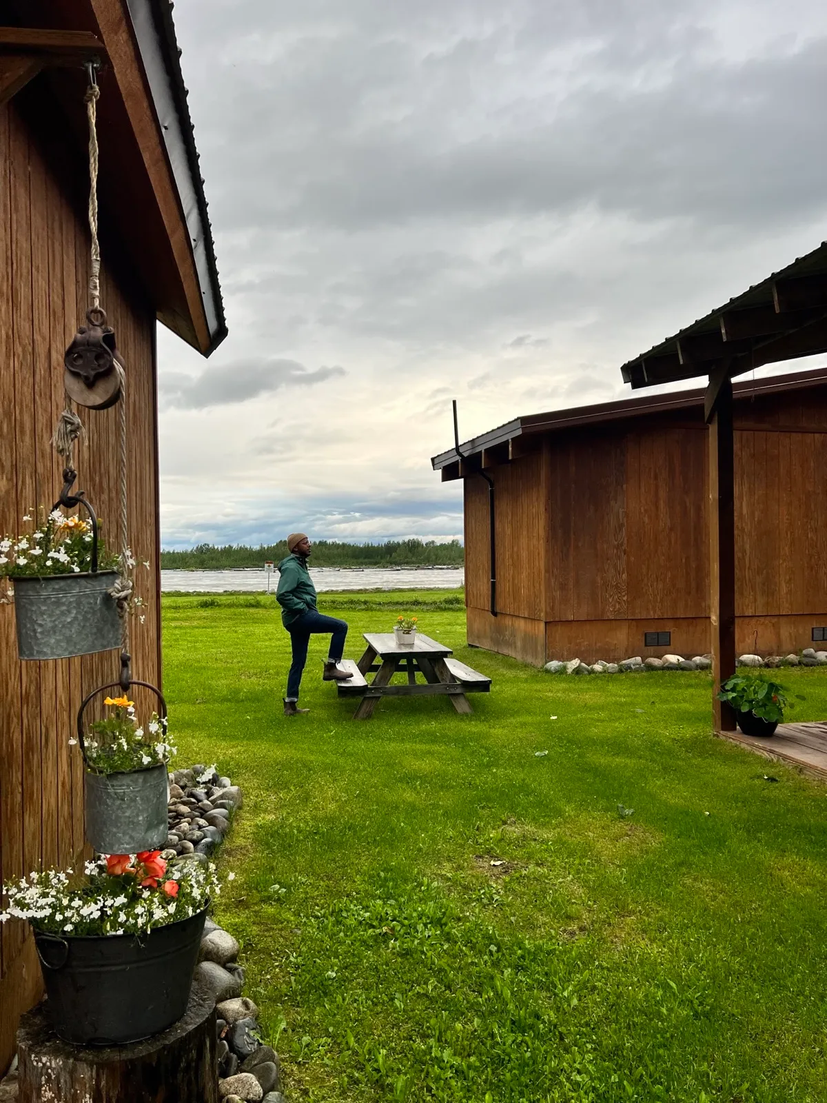 A wooden cabin porch with hanging flower buckets and a man in a beanie standing near a picnic table looking out at the river.